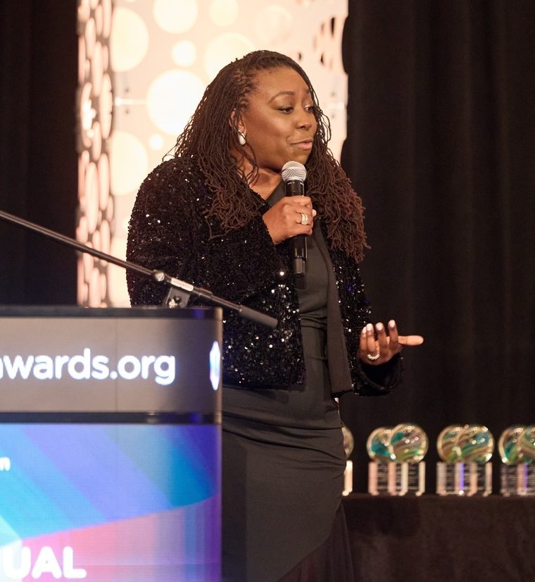 Brittany Davis Morris speaking on stage at the awards gala, with crystal trophies behind her
